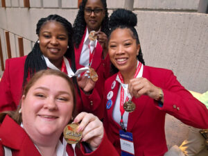 Four women wearing red jackets holding gold medals.