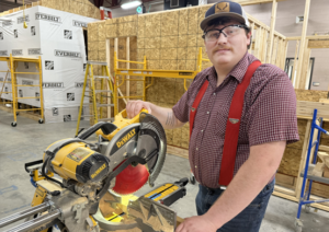 Construction Management: Construction student stands beside a miter saw in a hands-on building lab.